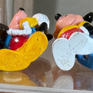 May include: Two vintage Disney character figurines, one featuring Donald Duck and the other Goofy. Donald Duck is wearing a blue shirt, red bow tie, and yellow shoes. Goofy is wearing a red shirt and blue pants. The figurines are marked "Applause" and "Hong Kong" and "China".