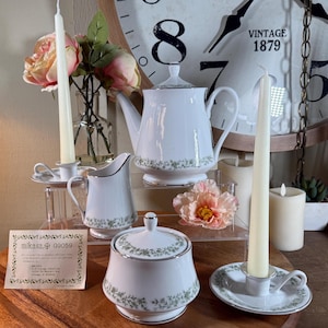 May include: A white Mikasa tea set with a floral pattern, including a teapot, creamer, sugar bowl, and candle holders. The set is displayed with candles, flowers, and a vintage clock in the background.