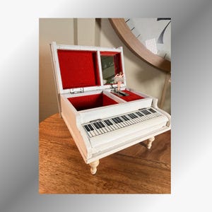 May include: A white piano-shaped jewelry box with a hinged lid, featuring a red velvet interior lining, a small rectangular mirror, and a miniature ballerina figurine that stands on a platform next to the keyboard keys on the top surface.
