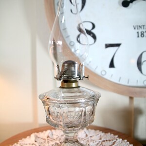 May include: A clear glass oil lamp with a decorative, faceted base and a round, clear glass chimney. The lamp is sitting on a white crocheted doily.