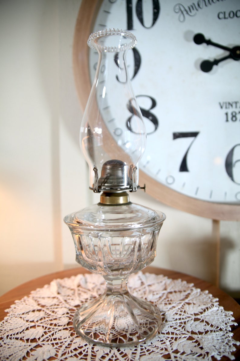 May include: A clear glass oil lamp with a decorative, faceted base and a round, clear glass chimney. The lamp is sitting on a white crocheted doily.