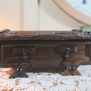 May include: A small, dark brown wooden chest with decorative metal accents. The chest has a rectangular shape with a hinged lid and two small drawers. It is supported by four decorative feet and is likely used for storing small items.