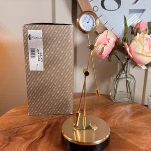 May include: A gold-toned Seiko desk clock with a round clock face and a unique, artistic design. The clock is displayed on a wooden surface, next to a box with the text "SEIKO OUF103G". The background includes a vase of pink roses.