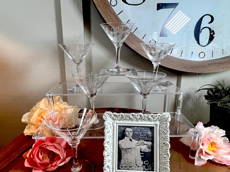 Op de afbeelding: Een set van zes heldere glazen martini glazen met gegraveerde ontwerpen worden gepresenteerd op een gelaagde acrylstandaard. De glazen zijn gerangschikt in een piramidevorm, met het grootste glas bovenaan en het kleinste glas onderaan. Op de voorgrond staat een ingelijste zwart-witfoto van een man in pak die een cocktailshaker vasthoudt. De foto heeft de tekst "Harry's of the New York Bar Paris - London - Cairo" en "ABC Mixing Cocktails".