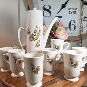 Könnte beinhalten: Ein cremefarbenes Vintage-Kaffee-Set mit floralem Design. Das Set umfasst eine hohe Kaffeekanne, mehrere Tassen und ein Serviertablett. Die Tassen und die Kaffeekanne haben eine gesprenkelte Oberfläche und ein Muster aus weißen Gänseblümchen und grünen Blättern.