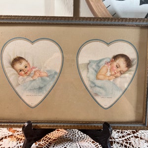 Puede incluir: Una impresión enmarcada de dos ilustraciones vintage de bebés durmiendo en marcos en forma de corazón. La impresión está en un marco dorado con un passe-partout marrón claro. El texto en la impresión dice "Sleep on in your little arms" y "Land of Nod".