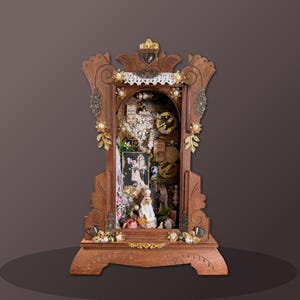 Könnte beinhalten: Ein hölzerner Schattenkasten mit kunstvollen Schnitzereien, gefüllt mit Vintage-Objekten, darunter ein Schwarz-Weiß-Foto, Uhrwerksteile und Blumenschmuck. Die Box ist mit goldfarbenen Verzierungen und Perlen verziert.