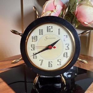 May include: Vintage Sessions clock in a ship's wheel design. The clock face is cream-colored with black numerals and hands, and the Sessions logo. The wheel is black with gold-colored spokes and a gold-colored base.