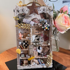 May include: A decorative shadow box with multiple shelves, filled with miniature objects. The box features a star and lace trim, with a variety of items including a doll, buttons, and printed ephemera. The color palette includes beige, gold, and touches of red.
