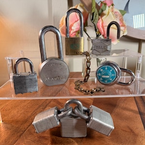 May include: A collection of various metal padlocks including a Master lock, a Yale lock, and a combination lock with a teal dial. The set features several silver-toned rectangular padlocks, a brass-toned padlock, and a metal chain connecting two locks.