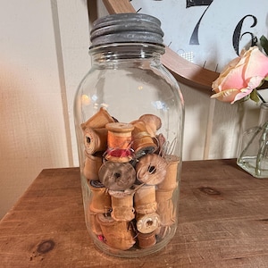 May include: A clear glass mason jar with a ribbed metal lid filled with various wooden thread spools. The spools are cylindrical and vary in light to dark brown tones, some featuring remnants of thread. The jar is transparent and cylindrical.