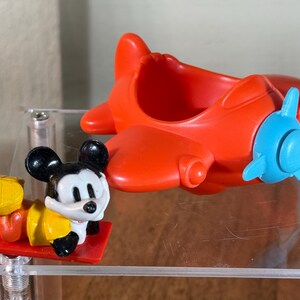 May include: A red toy airplane with a blue propeller and a Mickey Mouse figurine. The Mickey Mouse figure is black, white, and yellow, lying on a red platform. The airplane is made of plastic.