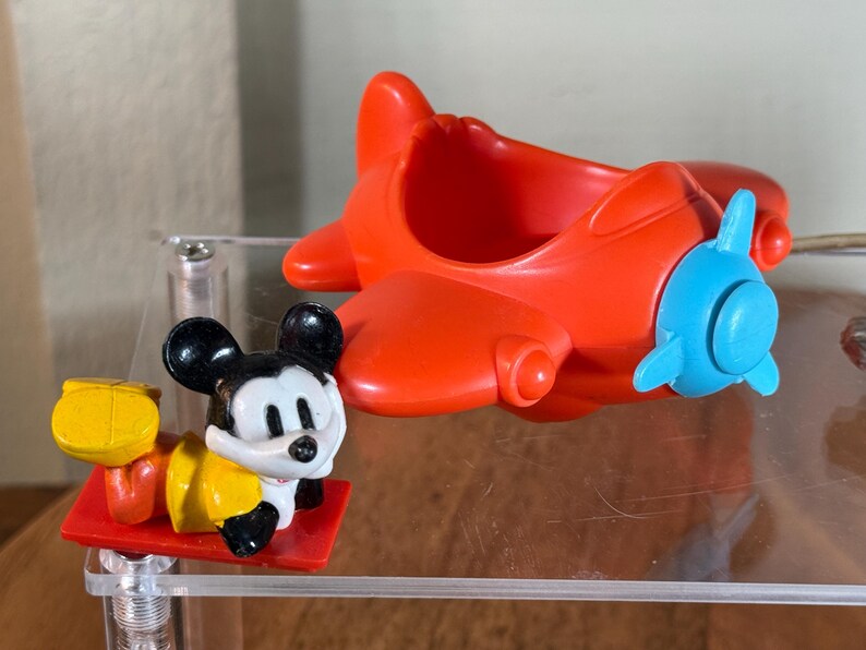 May include: A red toy airplane with a blue propeller and a Mickey Mouse figurine. The Mickey Mouse figure is black, white, and yellow, lying on a red platform. The airplane is made of plastic.