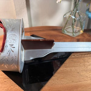 Puede incluir: Un visor de estereoscopio plateado vintage con un panel de visualización marrón oscuro. El visor tiene grabados florales decorativos y un cojín de terciopelo rojo. El estereoscopio está sobre una superficie negra.