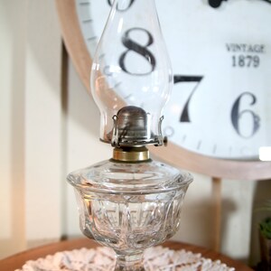 May include: A clear glass oil lamp with a round, textured base and a clear glass chimney. The lamp is sitting on a white lace doily.