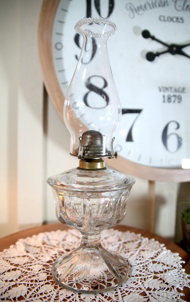 May include: A clear glass oil lamp with a round, textured base and a clear glass chimney. The lamp is sitting on a white lace doily.