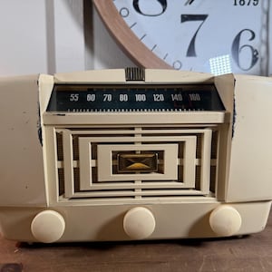 May include: Cream-coloured vintage radio with a rectangular speaker grille and three round knobs. The radio dial displays numbers from 55 to 160. The radio has a retro design, suggesting it's from the mid-20th century.