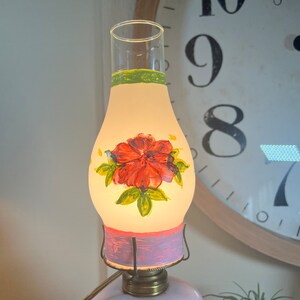 May include: A vintage oil lamp with a hand-painted floral design. The lamp has a clear glass shade with a pink and green painted band. The shade features a red flower with green leaves. The lamp base is a pale pink glass.
