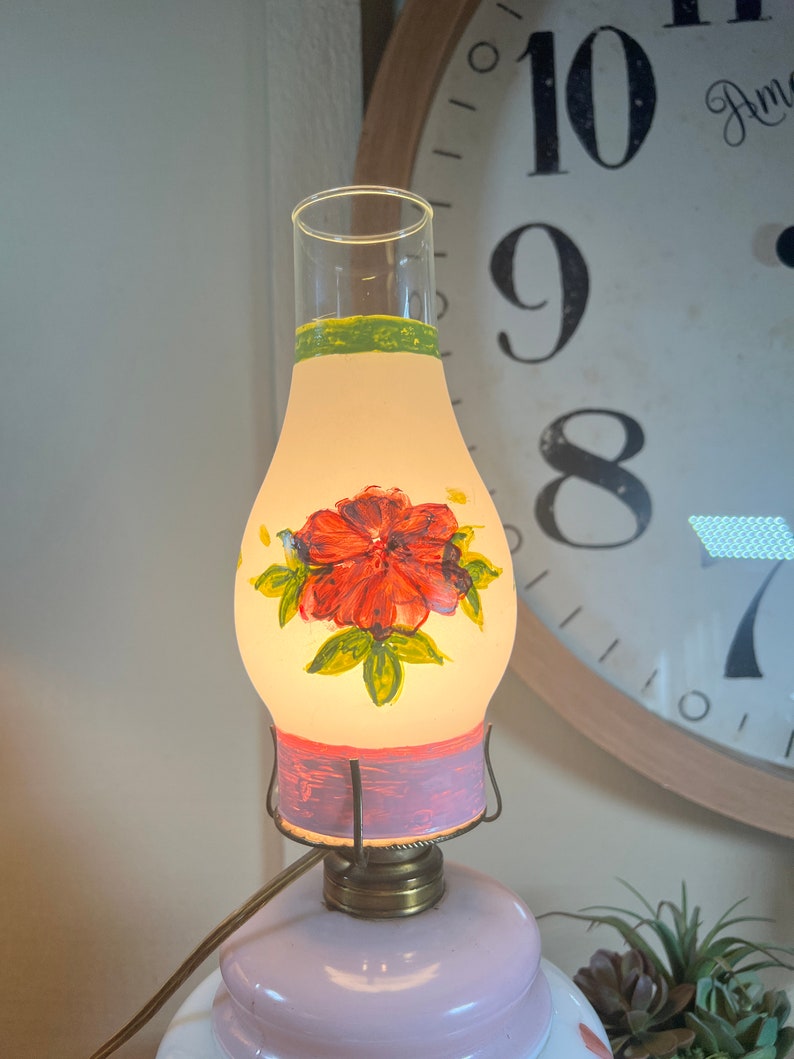 May include: A vintage oil lamp with a hand-painted floral design. The lamp has a clear glass shade with a pink and green painted band. The shade features a red flower with green leaves. The lamp base is a pale pink glass.