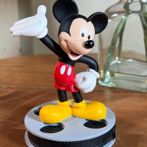May include: A Mickey Mouse figurine with black ears, a black body, red shorts, and yellow shoes. The figure has white gloves and is standing on a film reel base. The figurine is a collectible item.