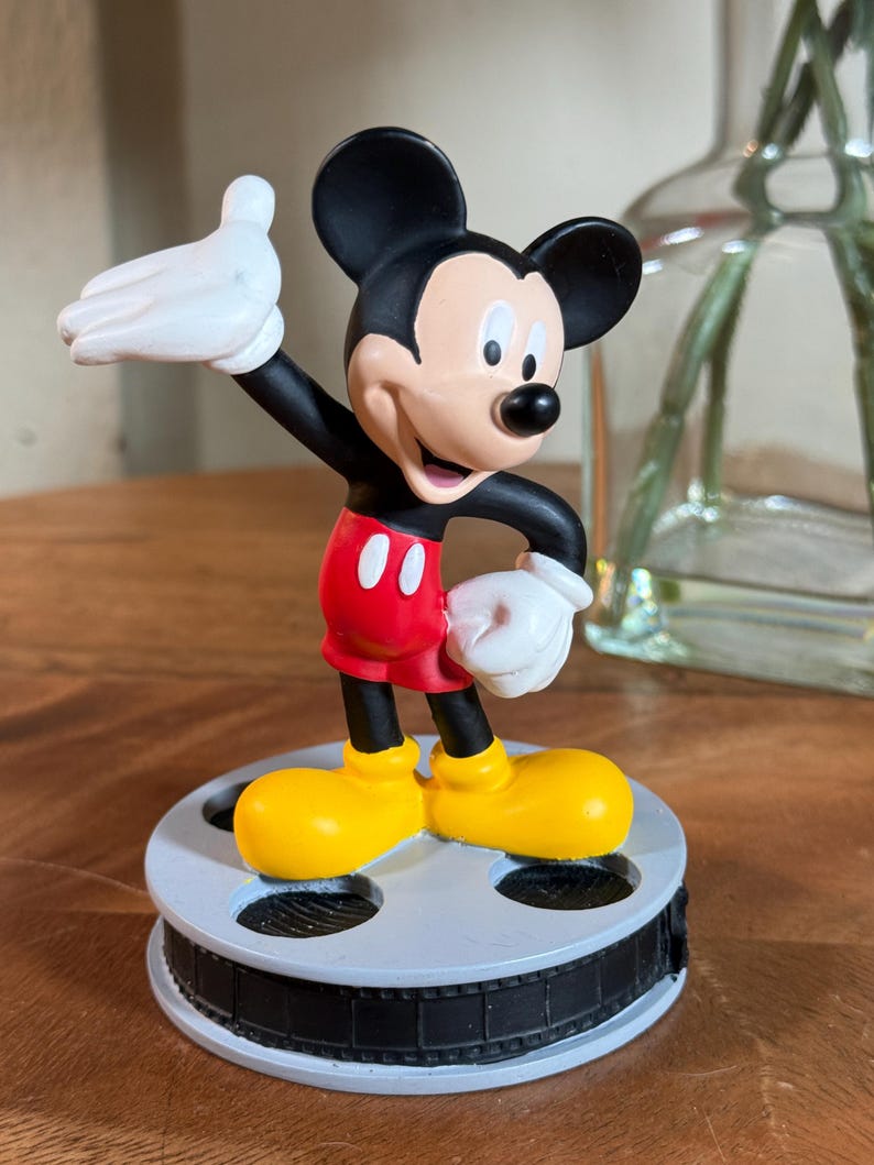 May include: A Mickey Mouse figurine with black ears, a black body, red shorts, and yellow shoes. The figure has white gloves and is standing on a film reel base. The figurine is a collectible item.