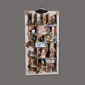 May include: A white, rectangular shadow box filled with various vintage items. The box has multiple compartments holding small figurines, a clock, a star, and other trinkets. Gold accents adorn the top corners. The items are arranged on a grey background.