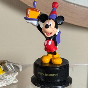 May include: A Mickey Mouse figurine holding a birthday cake with a lit candle. Mickey is wearing a party hat, red shorts, and yellow shoes. The base of the figurine reads "HAPPY BIRTHDAY!"