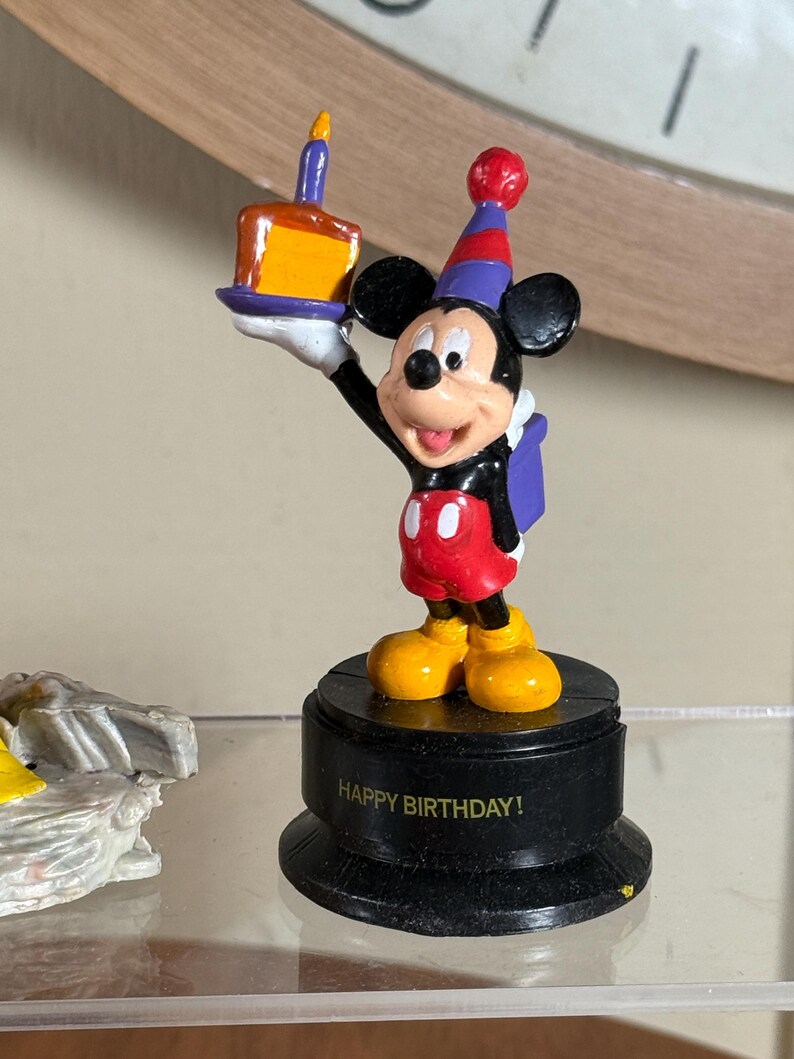 May include: A Mickey Mouse figurine holding a birthday cake with a lit candle. Mickey is wearing a party hat, red shorts, and yellow shoes. The base of the figurine reads "HAPPY BIRTHDAY!"