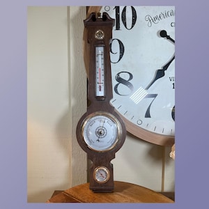 Puede incluir: Un barómetro de pared de madera con un termómetro y un higrómetro. El barómetro tiene una cara redonda con las palabras "Lluvia" y "Bueno" impresas en él. El termómetro es un tubo largo y delgado con un líquido rojo en su interior. El higrómetro es un pequeño dial redondo con una aguja que apunta al nivel de humedad.