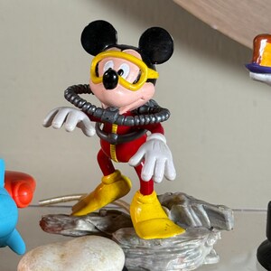 May include: A Mickey Mouse figurine in a scuba diving outfit. The figure is wearing a red diving suit, yellow flippers, and a yellow diving mask. The figure is standing on a gray rock base.