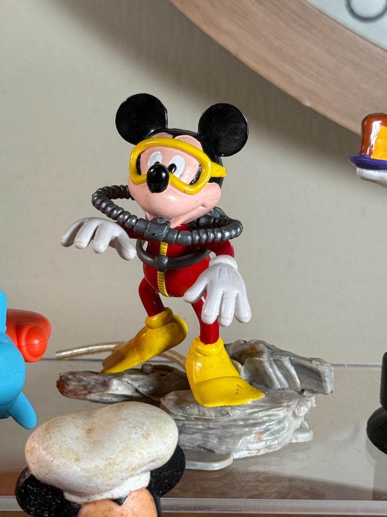 May include: A Mickey Mouse figurine in a scuba diving outfit. The figure is wearing a red diving suit, yellow flippers, and a yellow diving mask. The figure is standing on a gray rock base.