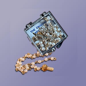 May include: A square, clear plastic case containing a white pearl necklace with gold-tone chain links. Inside the case, a gold-tone metal charm shaped like cat-eye glasses rests on the pearls. The text THE ORIGINAL is printed on the case interior.