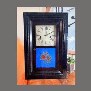 Könnte beinhalten: Antike Holzuhr mit schwarzem Rahmen und weißem Zifferblatt mit römischen Ziffern. Die Uhr hat einen blauen Hintergrund mit einem gemalten Rosenmuster.