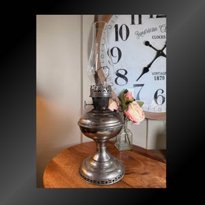 May include: Antique silver-colored oil lamp with a clear glass chimney. The lamp has a round base and a decorative design around the bottom. The background includes a wooden clock with the words "American Clocks" and "Vintage 1879".