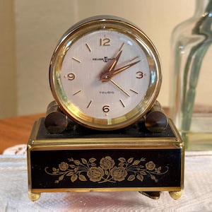 May include: A Reuge music table clock with a round gold-tone frame and a white textured dial featuring gold numerals at 3, 6, 9, and 12. The base is a rectangular black box with gold floral rose engravings and four small gold feet.
