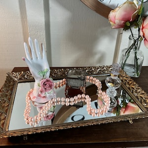 May include: A gold mirrored tray with ornate detailing, featuring a porcelain hand ring holder with pink roses, a small decorative box, and a strand of pink pearls.