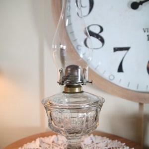 May include: A clear glass oil lamp with a cut glass base and a round, clear glass chimney. The lamp is sitting on a white lace doily on a wooden table.