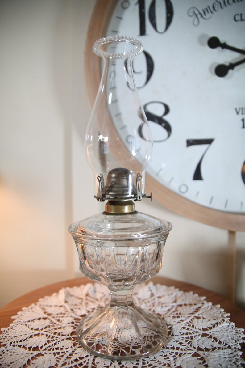 May include: A clear glass oil lamp with a cut glass base and a round, clear glass chimney. The lamp is sitting on a white lace doily on a wooden table.