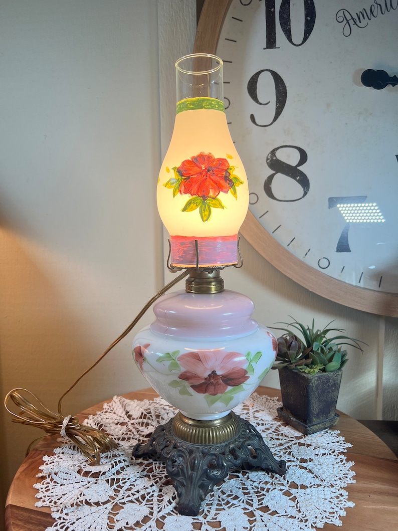 May include: A vintage electric lamp with a white and pink glass base and a clear glass shade with a painted floral design. The lamp has a black metal base with ornate details.