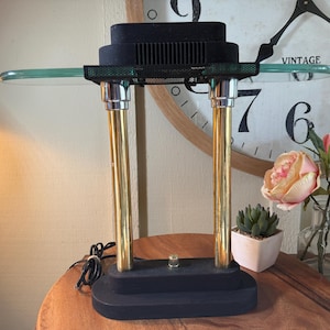 Può includere: Una lampada da scrivania nera e oro con base rettangolare e due supporti verticali color oro. La lampada ha un ripiano in vetro trasparente e una parte superiore nera con ventilazione. La lampada è alta circa 46 cm.
