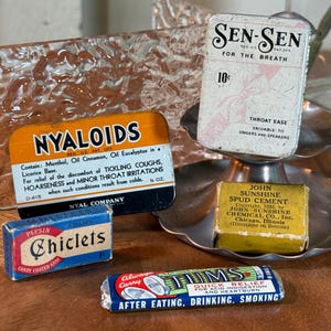 May include: A collection of vintage medicinal products, including Sen-Sen breath freshener, Nyaloids cough drops, Chiclets gum, John Sunshine Spud Cement, and Tums antacid tablets. The items are displayed on a wooden surface.