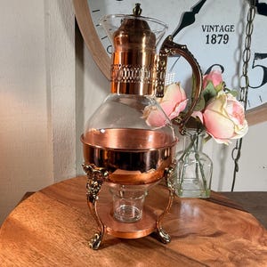 May include: A glass carafe with a copper-colored metal base and lid, featuring an ornate handle and a three-legged stand with decorative feet. The carafe has a clear glass body and a metallic collar with a perforated design, resting on a circular base.