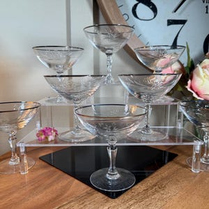 May include: A collection of clear champagne coupe glasses with silver rims and etched floral designs. The glasses are arranged on a tiered display, showcasing their elegant shape and delicate details. A vintage clock and flowers are in the background.