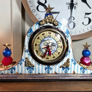 Pode incluir: Um relógio caprichoso com uma base listrada azul e branca e um mostrador com uma lua de desenho animado com um olho piscando. O relógio tem detalhes dourados e duas peças decorativas azuis e vermelhas.
