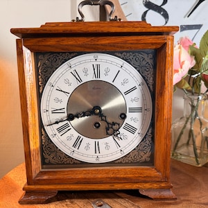 Könnte beinhalten: Eine quadratische Holzuhr mit hellbraunem Finish. Das Zifferblatt ist weiß mit schwarzen römischen Ziffern und verzierten schwarzen Details. Die Uhr hat eine silberfarbene Mitte und einen Griff oben.