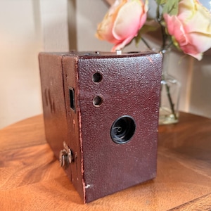 May include: A vintage, dark burgundy box camera with a textured surface. The camera has a circular lens, two small circular holes, and a rectangular viewfinder. The camera is a classic piece of photography equipment.