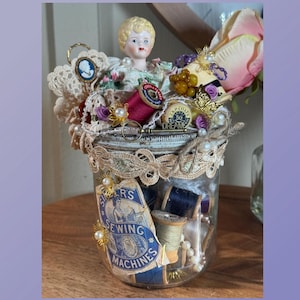 Puede incluir: Un tarro de cristal decorativo lleno de artículos de costura, incluyendo carretes de hilo, encaje y una cabeza de muñeca vintage. El tarro está adornado con perlas, un camafeo y una mariposa. La etiqueta dice "Singer's Sewing Machines."