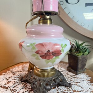 May include: A vintage white glass oil lamp with a pink base and a hand-painted floral design. The lamp has a brass base with a decorative scrollwork design and three legs.