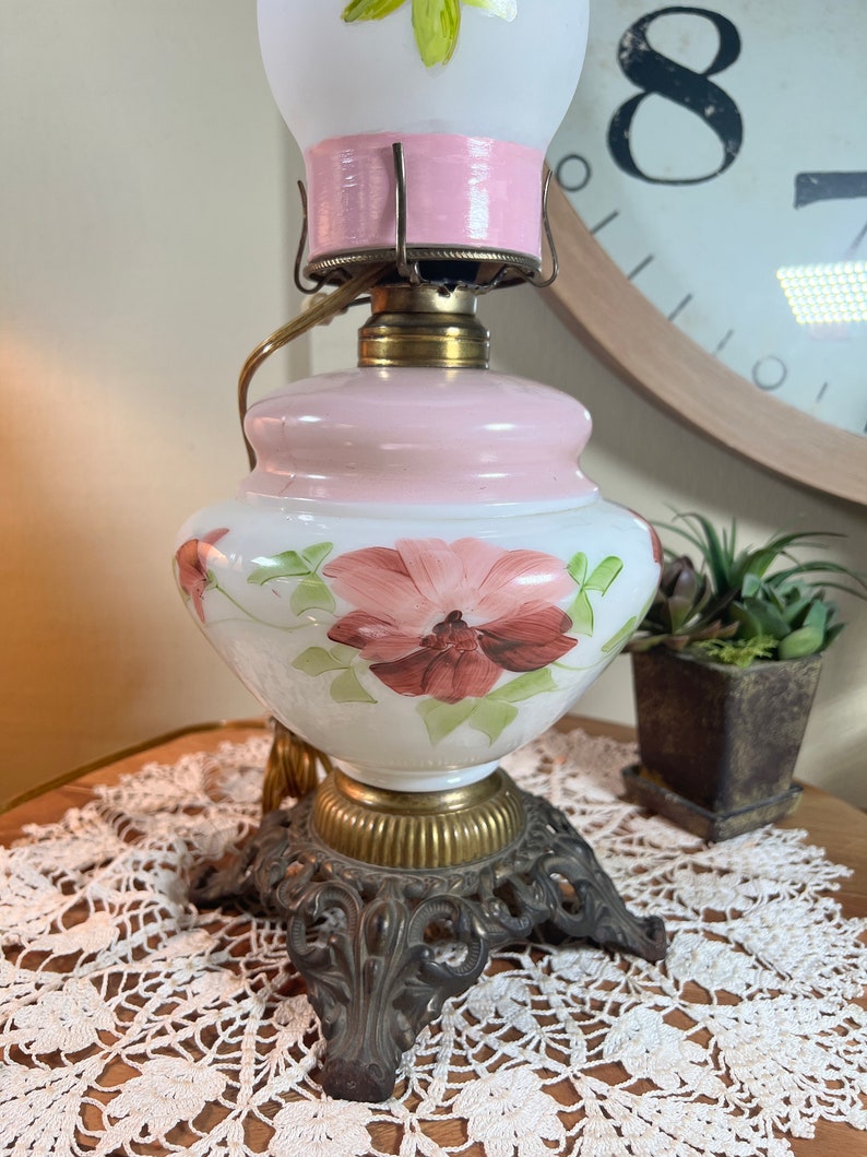 May include: A vintage white glass oil lamp with a pink base and a hand-painted floral design. The lamp has a brass base with a decorative scrollwork design and three legs.