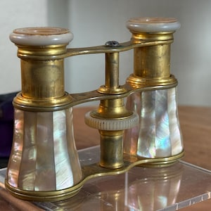 May include: Antique opera glasses with a brass frame and mother-of-pearl accents. The binoculars have a central focus wheel and white accents on the eyepieces. The design is a classic example of vintage optics.